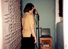 Here is a picture of Veronica 
singing in the vocal booth. As you look through the doorway, you see the
padding on the walls, Veronica standing at the microphone with headphones
on, and the powerbook running adatTalk.